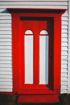 A wooden door with a symmetrical design is painted in a vibrant red and white color scheme. The door is set within a white exterior wall with horizontal siding.