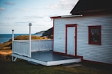 A small wooden house with white siding and red trim is situated in a coastal landscape. The house has a porch and overlooks an expanse of grassy land leading to the ocean. A cliff is visible in the background under a partly cloudy sky.