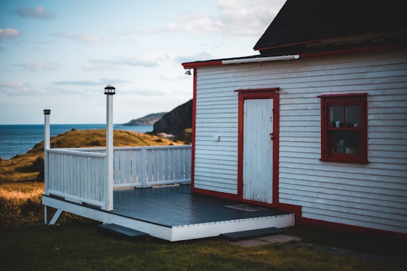 A small wooden house with white siding and red trim is situated in a coastal landscape. The house has a porch and overlooks an expanse of grassy land leading to the ocean. A cliff is visible in the background under a partly cloudy sky.