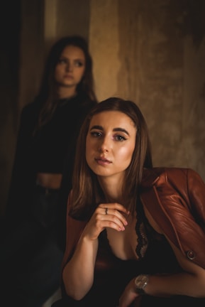 Two women are seen in a dimly lit setting with muted, warm tones. The woman in the foreground is wearing a brown leather jacket and a black top, looking directly into the camera with a serious expression. The background is out of focus, but another woman with long dark hair can be seen standing. The overall ambiance is moody and contemplative.
