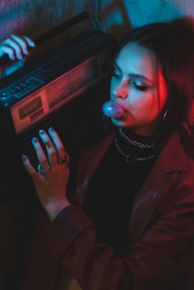 A person is holding a vintage boombox on their shoulder, blowing a bubble with gum. The scene is lit with red and blue neon lighting, creating a retro and edgy atmosphere. The person is wearing hoop earrings and rings, adding to the stylish look.