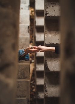 Two hands reaching towards each other between concrete walls, creating a sense of connection amidst a narrow, textured space.