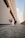 A small dog wearing a stylish white and black outfit with a red hoodie is standing confidently on a concrete pathway between two tall buildings. The urban setting includes modern architecture with clean lines and a soft blue sky in the background.