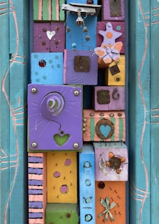 A vibrant, artistic assemblage featuring colorful wooden blocks adorned with various decorative elements such as a wire sculpture, butterfly shapes, heart cutouts, and metallic embellishments. The blocks are painted in a mix of colors including purple, pink, blue, and yellow, creating a playful and whimsical mosaic. The arrangement includes patterns like polka dots, stripes, and abstract designs, enhancing the visual complexity.