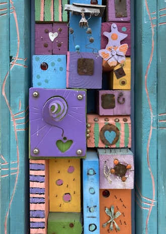 A vibrant, artistic assemblage featuring colorful wooden blocks adorned with various decorative elements such as a wire sculpture, butterfly shapes, heart cutouts, and metallic embellishments. The blocks are painted in a mix of colors including purple, pink, blue, and yellow, creating a playful and whimsical mosaic. The arrangement includes patterns like polka dots, stripes, and abstract designs, enhancing the visual complexity.