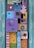 A vibrant, artistic assemblage featuring colorful wooden blocks adorned with various decorative elements such as a wire sculpture, butterfly shapes, heart cutouts, and metallic embellishments. The blocks are painted in a mix of colors including purple, pink, blue, and yellow, creating a playful and whimsical mosaic. The arrangement includes patterns like polka dots, stripes, and abstract designs, enhancing the visual complexity.