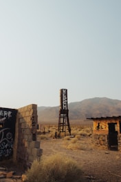 A desolate, desert landscape featuring an old water tower with the word 'MERSH' written on it. Surrounding the tower are dilapidated structures, one partially collapsed with graffiti on its walls. The terrain is dusty with sparse, dry vegetation, set against a backdrop of arid mountains under a clear sky.