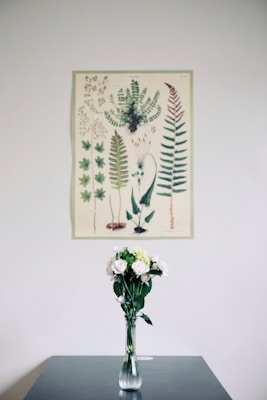 A minimalist and serene interior scene features a botanical illustration of various green ferns and leaves hanging on a white wall. Below the illustration, a clear glass vase holds a bouquet of white and light green flowers, creating a harmonious and natural aesthetic.