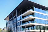 A modern, multi-story office building with a large overhanging roof and extensive glass windows. The architecture features clean lines, flat surfaces, and a combination of steel, glass, and concrete elements. Nearby, there are some trees and greenery adding a touch of nature to the urban setting.