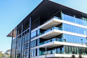 A modern, multi-story office building with a large overhanging roof and extensive glass windows. The architecture features clean lines, flat surfaces, and a combination of steel, glass, and concrete elements. Nearby, there are some trees and greenery adding a touch of nature to the urban setting.