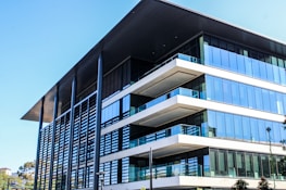 A modern, multi-story office building with a large overhanging roof and extensive glass windows. The architecture features clean lines, flat surfaces, and a combination of steel, glass, and concrete elements. Nearby, there are some trees and greenery adding a touch of nature to the urban setting.