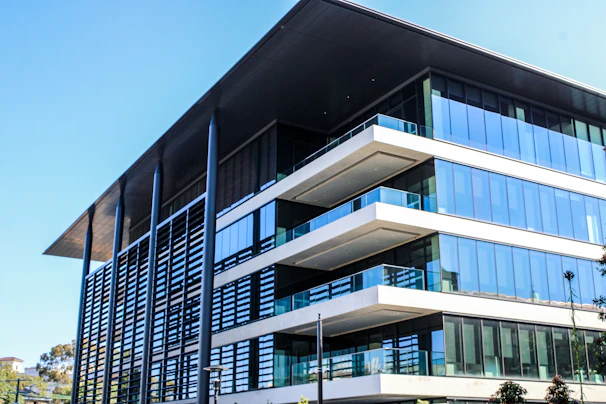 A modern, multi-story office building with a large overhanging roof and extensive glass windows. The architecture features clean lines, flat surfaces, and a combination of steel, glass, and concrete elements. Nearby, there are some trees and greenery adding a touch of nature to the urban setting.