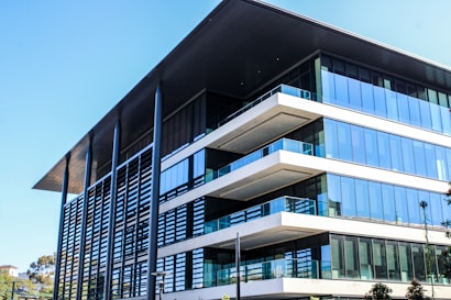 A modern, multi-story office building with a large overhanging roof and extensive glass windows. The architecture features clean lines, flat surfaces, and a combination of steel, glass, and concrete elements. Nearby, there are some trees and greenery adding a touch of nature to the urban setting.