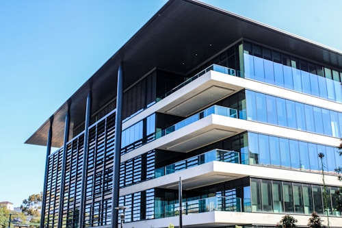 A modern, multi-story office building with a large overhanging roof and extensive glass windows. The architecture features clean lines, flat surfaces, and a combination of steel, glass, and concrete elements. Nearby, there are some trees and greenery adding a touch of nature to the urban setting.
