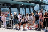 A group of people gathers in front of a rail or barrier, some with backpacks and summer clothing, suggesting a warm day. They seem to be waiting or queuing for an event or attraction. Behind them, signage for day tours and a modern building structure are visible.