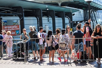 A group of people gathers in front of a rail or barrier, some with backpacks and summer clothing, suggesting a warm day. They seem to be waiting or queuing for an event or attraction. Behind them, signage for day tours and a modern building structure are visible.