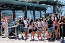 A group of people gathers in front of a rail or barrier, some with backpacks and summer clothing, suggesting a warm day. They seem to be waiting or queuing for an event or attraction. Behind them, signage for day tours and a modern building structure are visible.