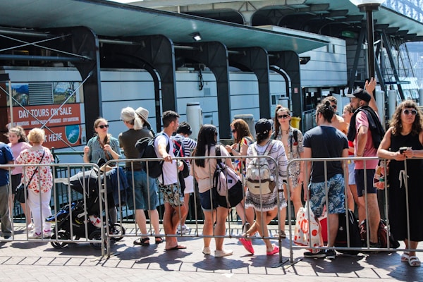 A group of people gathers in front of a rail or barrier, some with backpacks and summer clothing, suggesting a warm day. They seem to be waiting or queuing for an event or attraction. Behind them, signage for day tours and a modern building structure are visible.