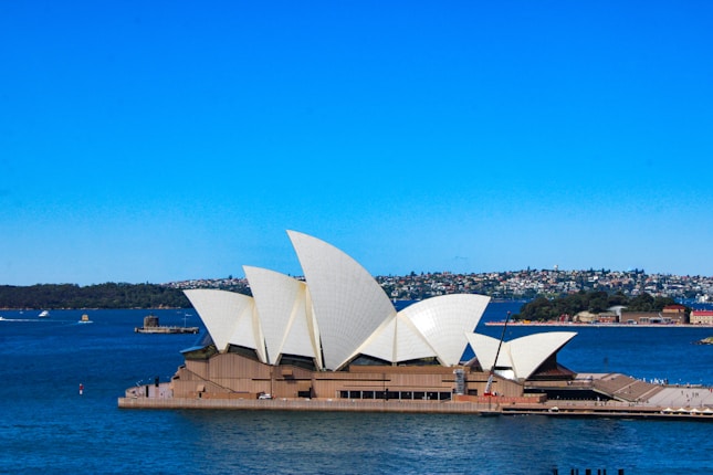 The iconic Sydney Opera House stands prominently against a bright blue sky and the expansive waters around it. The building's distinctive white shell-like structures are contrasted with the calm blue of the surrounding harbor. In the background, a shoreline with green hills and scattered buildings is visible.