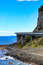 A coastal bridge stretches over rocky terrain with the ocean visible in the background. The structure is elevated, supported by large pillars, and the landscape includes lush greenery and a winding shoreline.