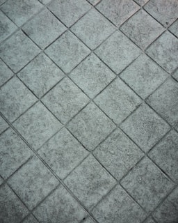 A textured, grey tiled surface with a repeated diamond pattern. The tiles appear to be made of stone or concrete and have a rough, weathered look.