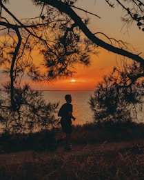 A silhouette of a person jogging along a path near a body of water at sunset. The sky is filled with vibrant hues of orange and yellow as the sun descends towards the horizon. Tree branches frame the scene, adding a natural border to the image.