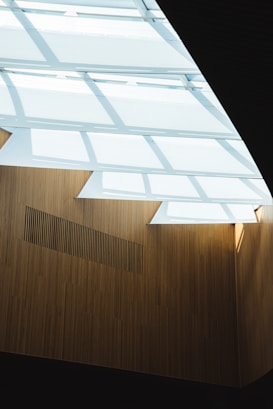 A modern architectural structure featuring large, diagonal beams allowing sunlight to penetrate through. The interior walls are clad in a vertical, wooden panel design, creating a warm contrast with the incoming light.