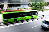 A bright green public bus with the text 'SG BUS' is parked on the side of a busy street. The bus has a red top and is adjacent to a Starbucks coffee shop. Several pedestrians are walking nearby, and there is a crossing with traffic lights visible. Another vehicle is passing by on the road.