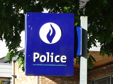 A blue sign with white lettering displaying the word 'Police' next to an emblem. The background shows leafy green foliage partially shading the sign, and parts of a building with a brick exterior are visible.
