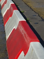 A row of red and white interlocking concrete barriers lines the edge of a paved area. The sunlight casts shadows on the ground, highlighting the texture of the barriers and the wet surface nearby.