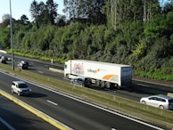 A large truck with branding on its trailer is traveling on a multi-lane highway surrounded by several cars. The highway is bordered by lush green trees and a clear blue sky is visible above.