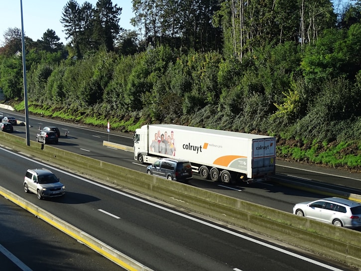 A large truck with branding on its trailer is traveling on a multi-lane highway surrounded by several cars. The highway is bordered by lush green trees and a clear blue sky is visible above.