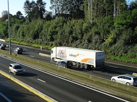 A large truck with branding on its trailer is traveling on a multi-lane highway surrounded by several cars. The highway is bordered by lush green trees and a clear blue sky is visible above.