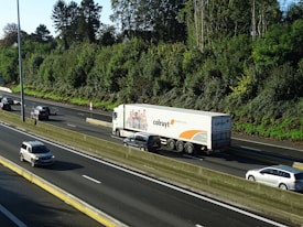 A large truck with branding on its trailer is traveling on a multi-lane highway surrounded by several cars. The highway is bordered by lush green trees and a clear blue sky is visible above.
