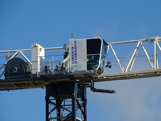 A close-up view of a tower crane's operator cabin, mounted on a horizontal beam. The cabin is mostly white with large windows, and the structure is labeled with the name 'Linden Comansa'. There is a blue sky in the background.