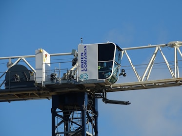 A close-up view of a tower crane's operator cabin, mounted on a horizontal beam. The cabin is mostly white with large windows, and the structure is labeled with the name 'Linden Comansa'. There is a blue sky in the background.