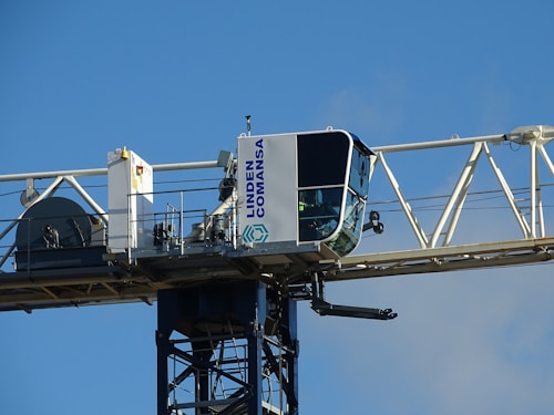 A close-up view of a tower crane's operator cabin, mounted on a horizontal beam. The cabin is mostly white with large windows, and the structure is labeled with the name 'Linden Comansa'. There is a blue sky in the background.