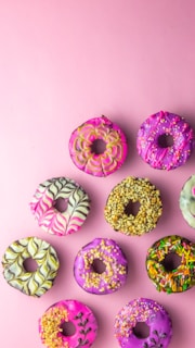 An arrangement of colorful donuts on a pink background, each with unique designs and toppings. The donuts feature a variety of icing patterns, including swirls, sprinkles, and crushed nuts.