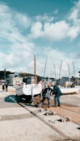 Several people are working together to move a white boat on a trailer along a harbor walkway. The background consists of a row of sailboats with tall masts and a cluster of quaint seaside buildings under a partly cloudy sky. The scene suggests a community effort at a busy harbor area.