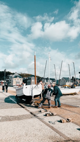 Several people are working together to move a white boat on a trailer along a harbor walkway. The background consists of a row of sailboats with tall masts and a cluster of quaint seaside buildings under a partly cloudy sky. The scene suggests a community effort at a busy harbor area.