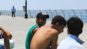 A group of young men are sitting on the edge of a seaside promenade. One of them is shirtless, while another wears a cap and a sleeveless shirt. They appear to be engaged in conversation. In the background, the ocean is visible along with a railing and a walkway. Further away, a man is strolling toward the camera.