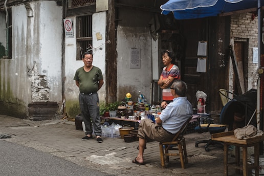 Three individuals are gathered in an outdoor setting next to a deteriorating building. A man in a green shirt stands with hands behind his back, another woman in a striped shirt is engaged in conversation, and a man wearing a blue shirt is seated on a chair. Various items like bottles and bowls are displayed on a table nearby, suggesting a small market or gathering. The surroundings include worn walls and a mixture of chairs.