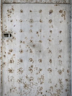 A rusty metal surface covered in numerous round patches of corrosion. The texture appears rough with a mixture of brown and orange oxide formations against a faded gray background. A visible rectangular latch or slot is present on the left side.