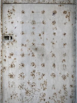 A rusty metal surface covered in numerous round patches of corrosion. The texture appears rough with a mixture of brown and orange oxide formations against a faded gray background. A visible rectangular latch or slot is present on the left side.