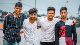 Four young men stand closely together with their arms around each other. They are outdoors, and the background shows a cloudy sky and some trees. Each person is smiling or looking cheerful.