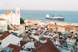 A scenic view of a coastal city with red-tiled rooftops and a historic white church overlooking the sea. A large cargo ship is visible on the calm waters, contrasting with the vibrant colors of the buildings and landscape.