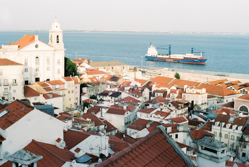 A scenic view of a coastal city with red-tiled rooftops and a historic white church overlooking the sea. A large cargo ship is visible on the calm waters, contrasting with the vibrant colors of the buildings and landscape.