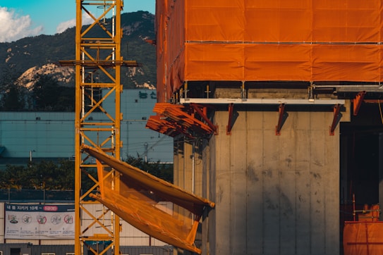 A section of a construction site with a partially built structure. The structure is covered in bright orange netting and scaffolding. In the foreground, yellow construction equipment and support beams are visible. The background features mountains and a building with a blue exterior. The sun casts long shadows, adding contrast to the scene.