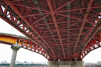 A detailed view of a large steel bridge structure with red and orange iron beams intersecting in a geometric pattern. The bridge is elevated, supported by concrete pillars, with a cityscape visible in the background.