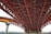 A detailed view of a large steel bridge structure with red and orange iron beams intersecting in a geometric pattern. The bridge is elevated, supported by concrete pillars, with a cityscape visible in the background.